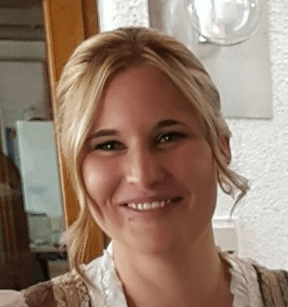 Michelle Auer, eine Frau mit blondem Haar, lächelt in einem Innenraum in die Kamera, wo sie vor einer weißen Wand und einem hölzernen Türrahmen steht.