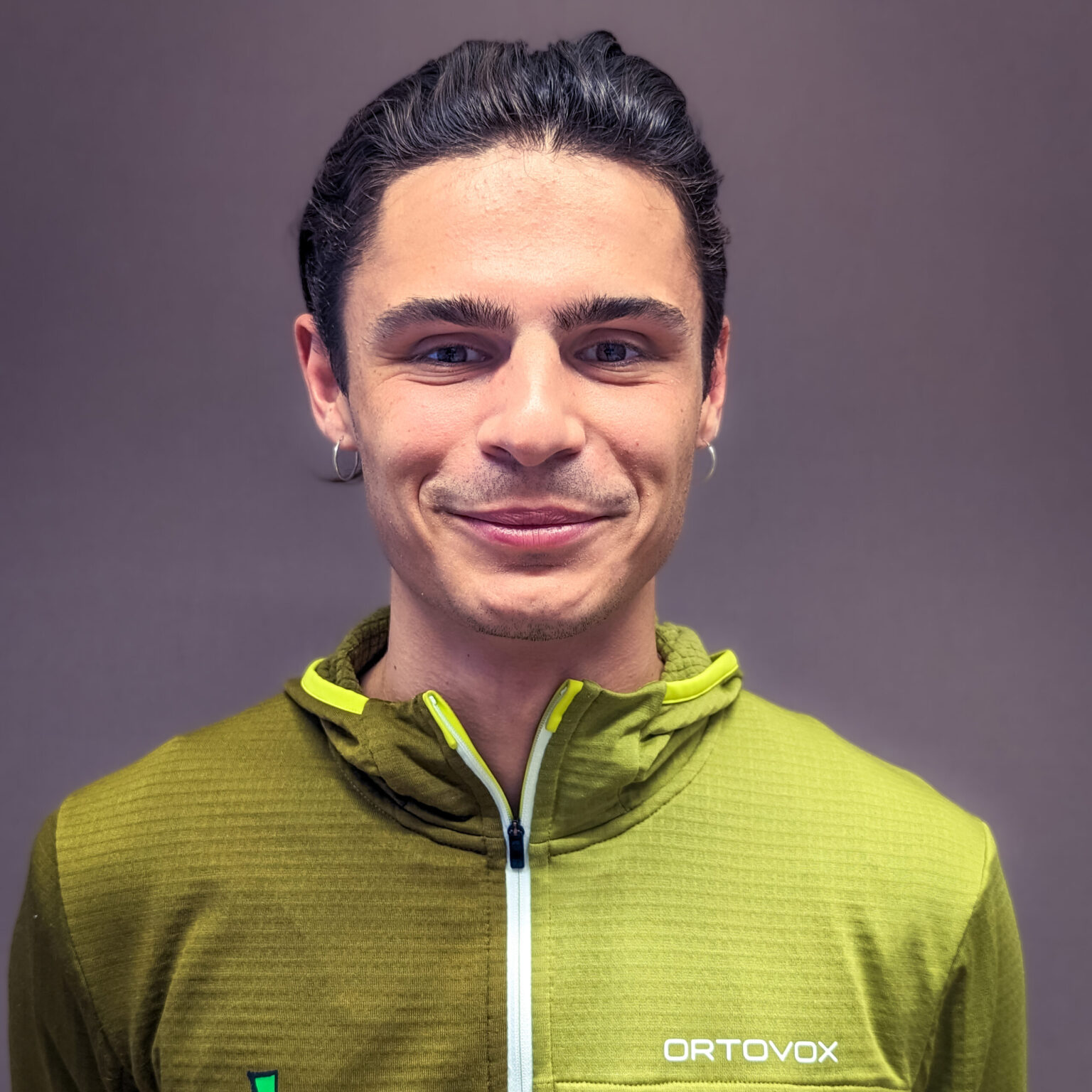 Felix Bäcker, ein junger Mann mit kurzen, dunklen Haaren und grüner Ortovox-Jacke, steht vor einem schlichten, neutralen Hintergrund, schaut in die Kamera und lächelt leicht.