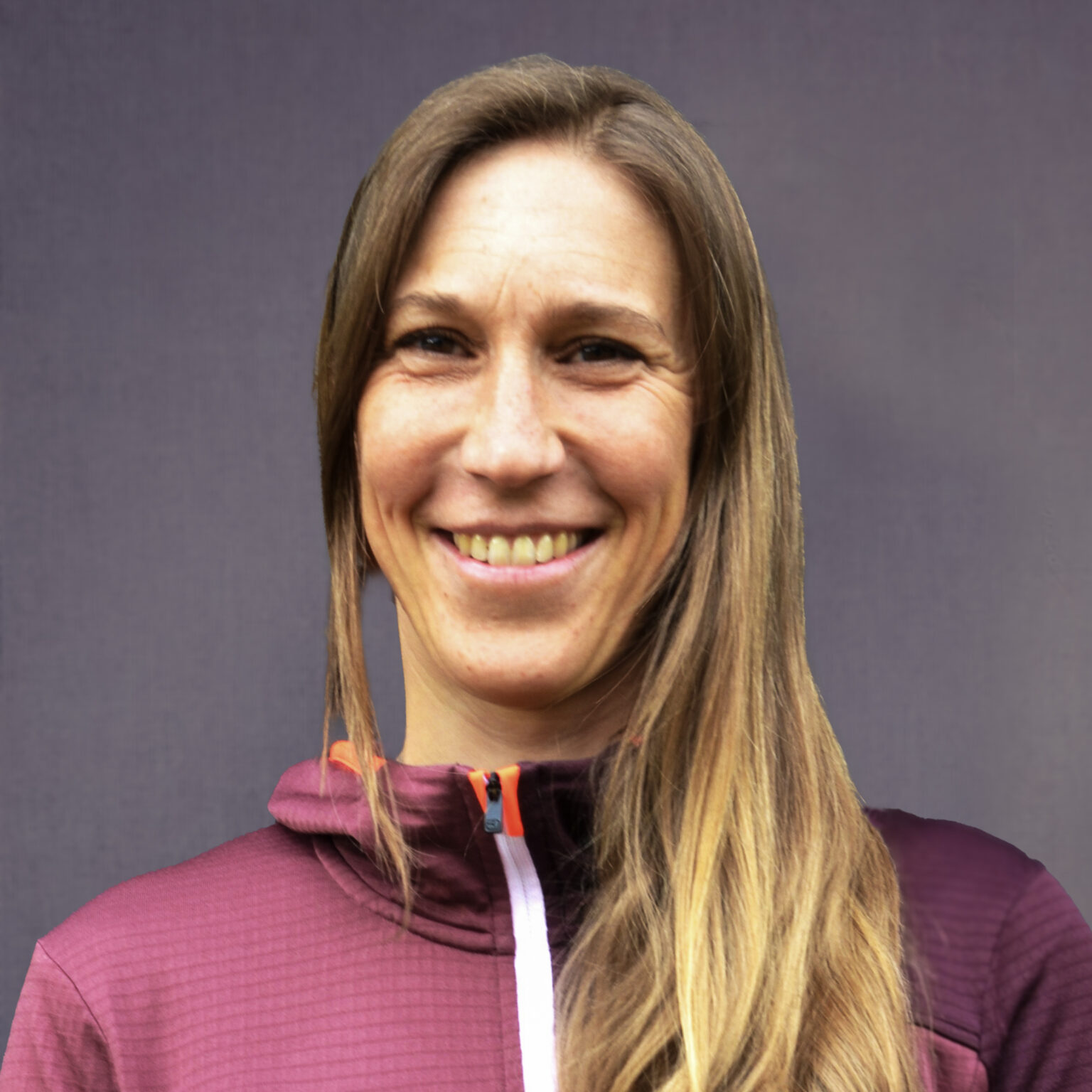 Sina Hölscher, eine Frau mit langen blonden Haaren, trägt eine kastanienbraune Jacke mit orangefarbenem Reißverschluss und lächelt in die Kamera vor einem schlichten, neutralen Hintergrund.