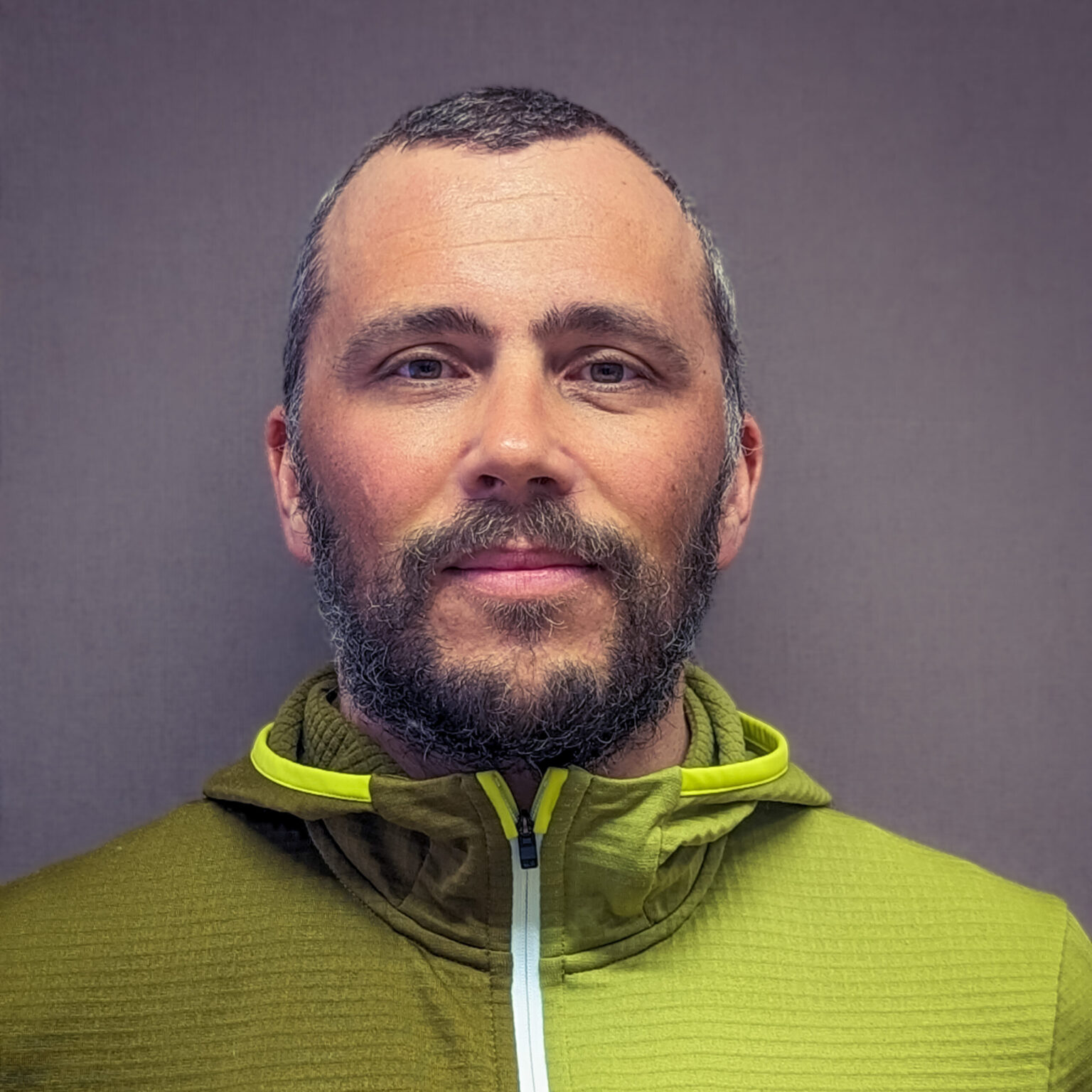 Sebastian Pilloni, ein Mann mit Bart und kurzen Haaren, steht vor einem schlichten grauen Hintergrund, trägt eine grüne Jacke mit Reißverschluss und schaut direkt in die Kamera.