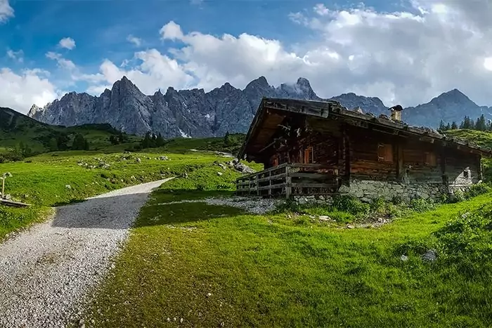 Eine Holzhütte steht auf einem grasbewachsenen Hügel neben einem Kiesweg, mit zerklüfteten Berggipfeln und einem teilweise bewölkten Himmel im Hintergrund - perfekt für landschaftliche Tapeten-Downloads.