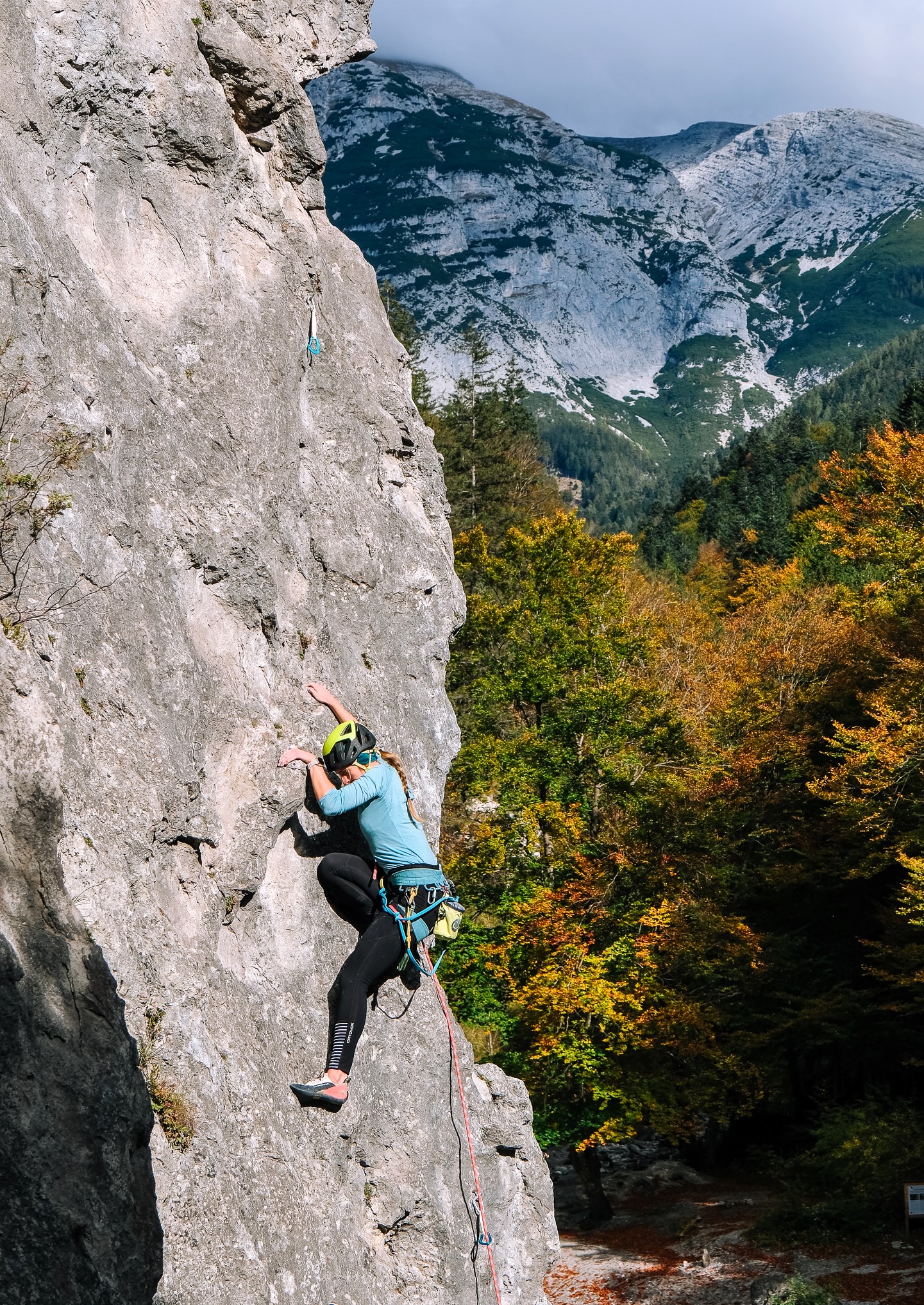 Eine Person in Kletterausrüstung erklimmt eine steile Felswand mit einem Berg und Bäumen im Hintergrund, eingefangen durch beeindruckende Foto-/ Filmaufnahmen & Drohnen für eine atemberaubende Perspektive.