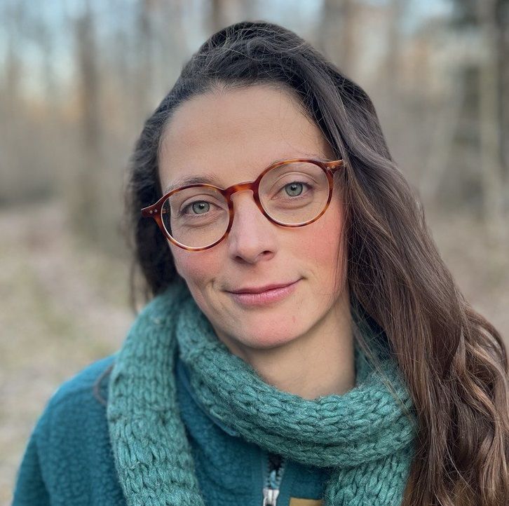 Nadine Werner, eine Frau mit langen braunen Haaren, trägt eine runde Brille, einen blaugrünen Strickschal und eine blaugrüne Fleecejacke, während sie im Freien steht und die Bäume im Hintergrund leicht verschwimmen.