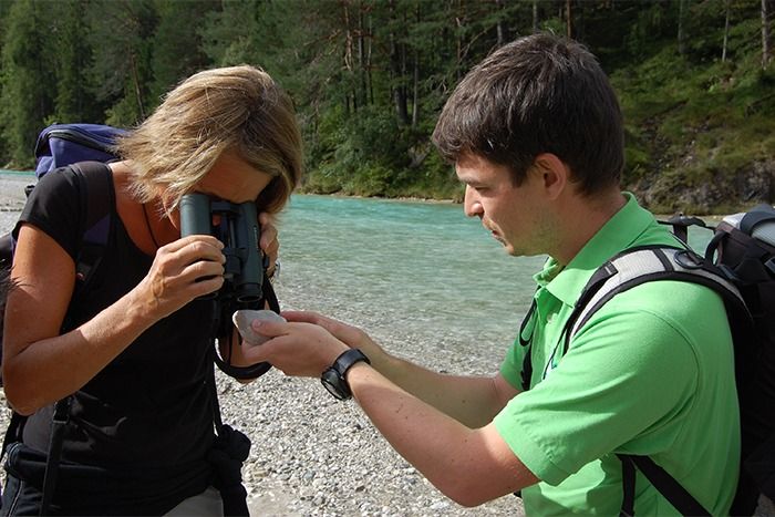 Eine Frau untersucht einen Gegenstand in der Hand eines Mannes mit einer Lupe an einem Flussufer, mit Bäumen und Wasser im Hintergrund, während sie über die letzten Downloads spricht.