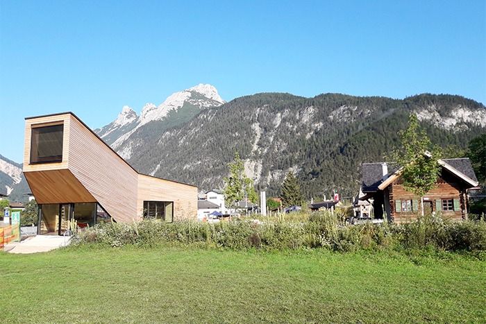 Ein modernes Holzgebäude mit abgewinkeltem Dach steht auf einer Wiese und bietet eine atemberaubende Aussicht und einfache Downloads, mit traditionellen Häusern und bewaldeten Bergen im Hintergrund unter einem klaren blauen Himmel.