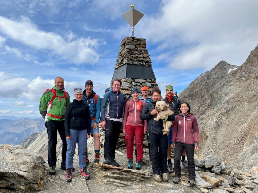 Eine Gruppe von zehn Personen und ein Hund stehen an einer steinernen Gipfelmarkierung auf einem Berg in den Tiroler Alpen, umgeben von felsigen Gipfeln unter einem teilweise bewölkten Himmel.