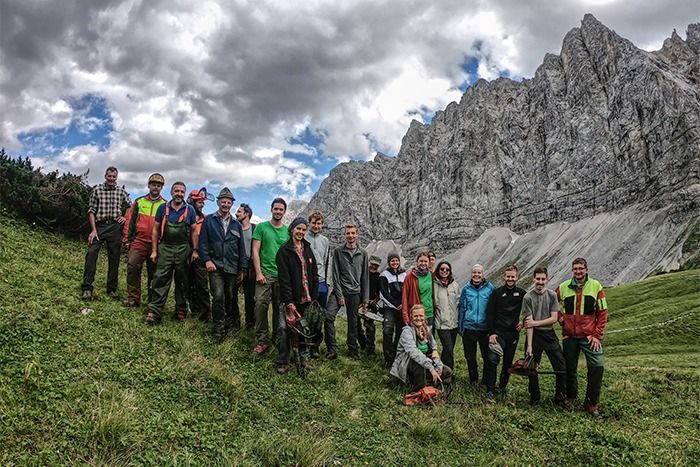 Eine Gruppe von Menschen posiert auf einem grasbewachsenen Hang mit felsigen Bergen und einem wolkenverhangenen Himmel im Hintergrund und hält einen Moment fest, der sich perfekt zum Teilen oder für zukünftige Downloads eignet.