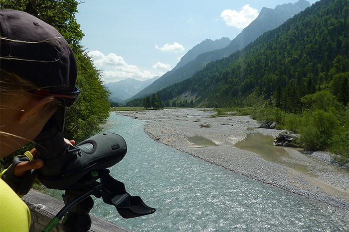 Eine Person beobachtet mit einem Spektiv einen sich schlängelnden Fluss, der von bewaldeten Bergen umgeben ist, unter einem teilweise bewölkten Himmel und hält die Ansicht als digitalen Download fest.