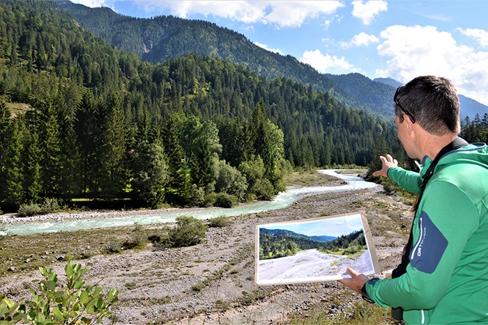Eine Person in grüner Jacke hält ein Foto der Landschaft in der Hand, während sie auf einen Fluss zeigt, der durch ein bewaldetes Bergtal fließt, und suggeriert Downloads von landschaftlichen Momenten, die man für immer in Erinnerung behalten wird.