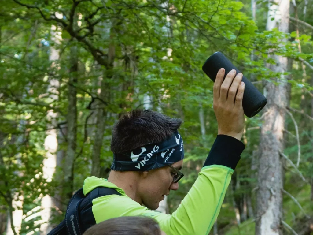 Eine Person, die eine Brille und ein Stirnband trägt, hält in einer Waldlandschaft einen schwarzen tragbaren Lautsprecher über den Kopf - vielleicht inspiriert durch die aktuelle Forschung über die Wirkung von Musik in der Natur.