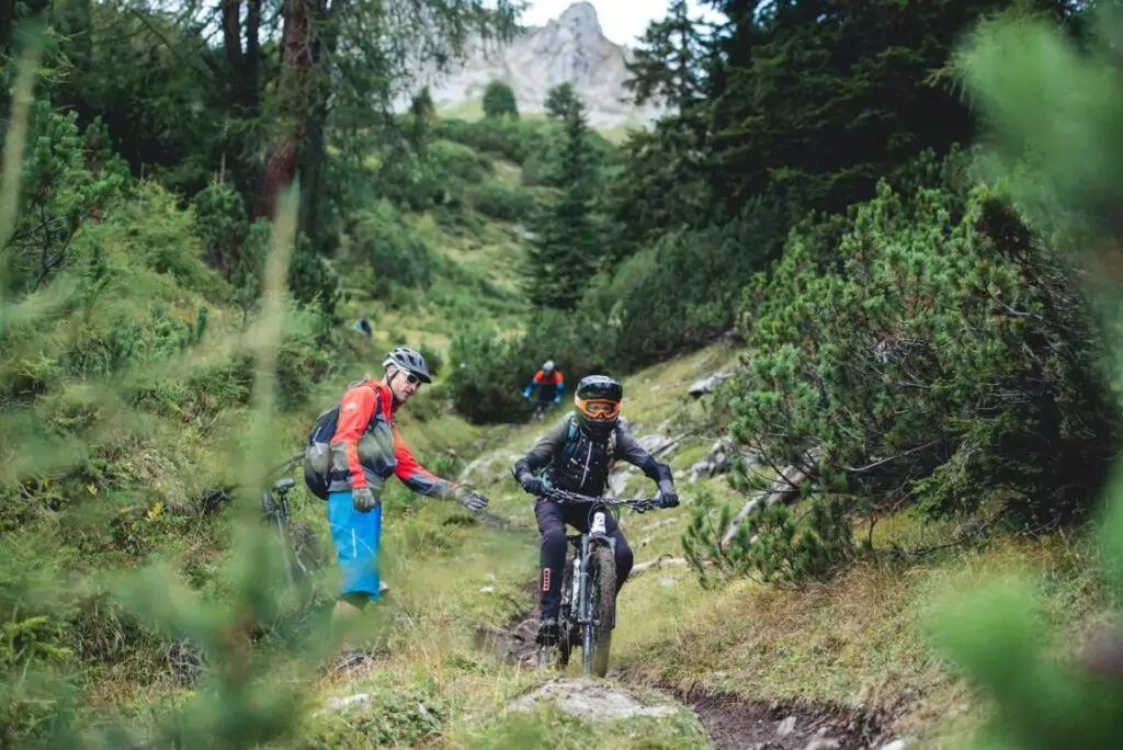 Zwei Mountainbiker genießen das Mountainbiken im Karwendel auf einem schmalen, felsigen Waldweg, umgeben von üppigem Grün und Bäumen, während im Hintergrund weitere Biker folgen.