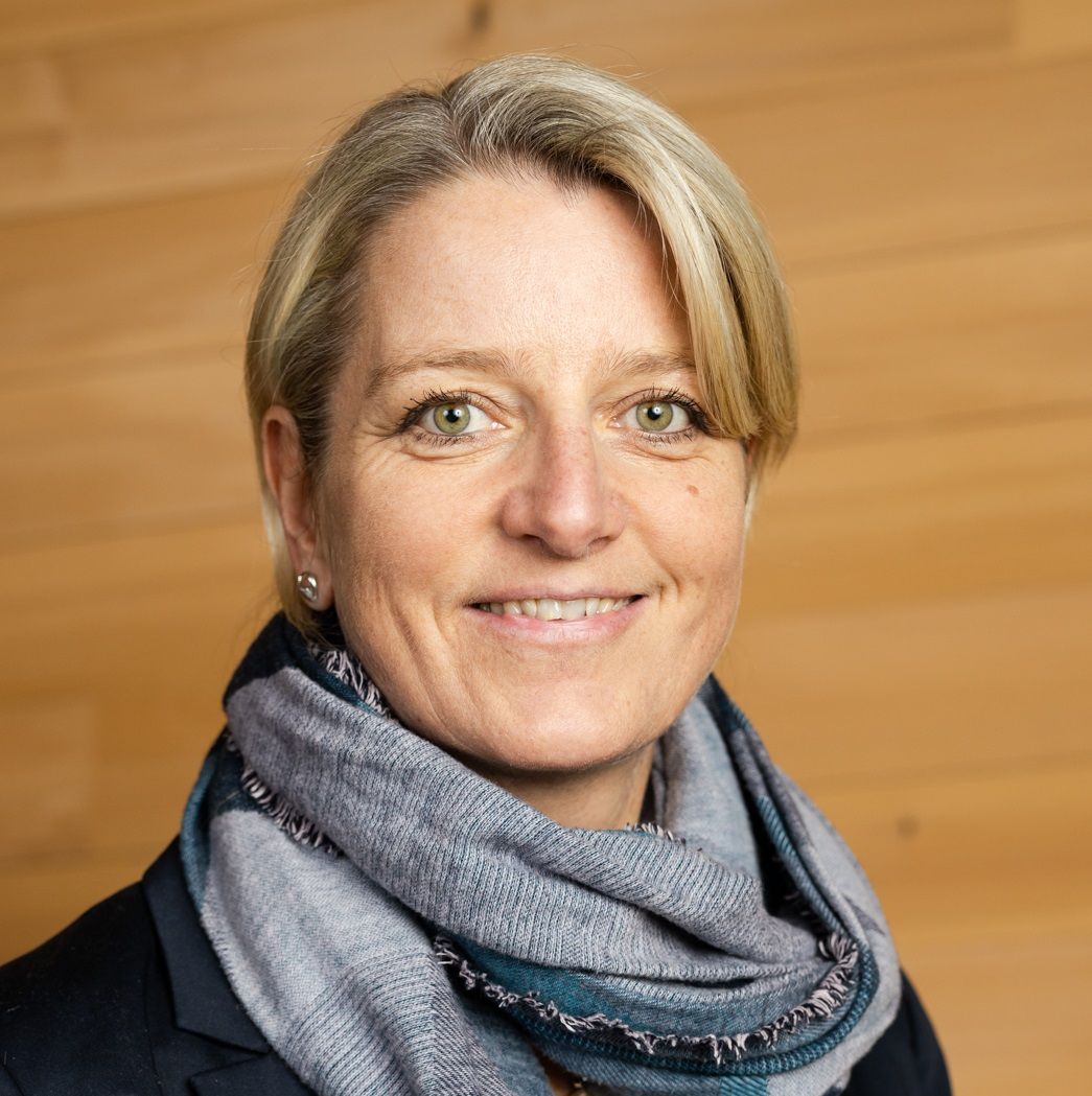 Sandra Rinner, eine Frau mit blondem Haar und grünen Augen, die einen dunklen Blazer und einen grauen Schal trägt, lächelt vor einem hölzernen Hintergrund.