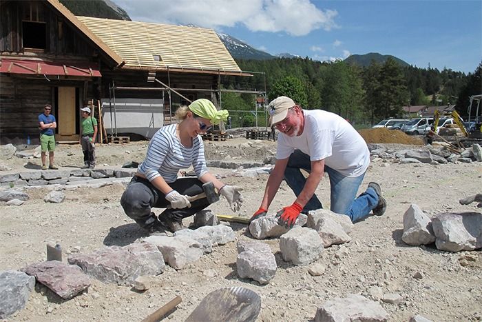 Zwei Personen mit Handschuhen arbeiten zusammen, um Steine auf einer Baustelle im Freien zu platzieren, mit einem Holzgebäude und Bergen im Hintergrund, während Downloads auf ihren Geräten ihnen den Weg weisen.
