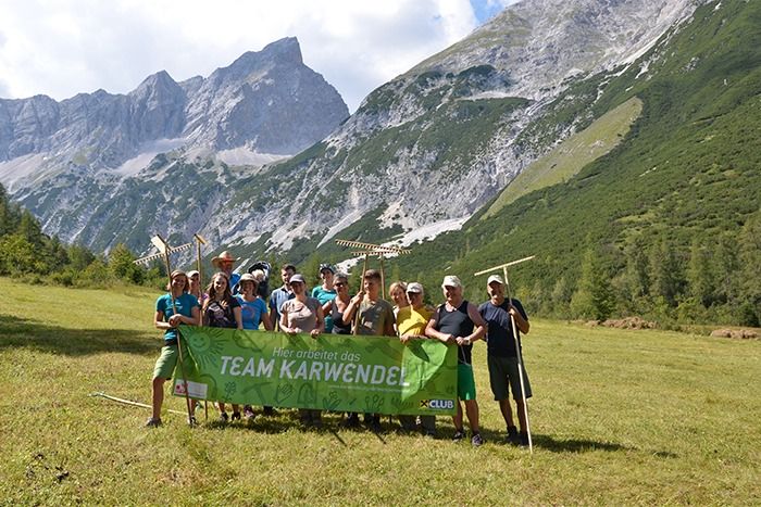 Eine Gruppe von Personen mit einem grünen "Team Karwendel"-Banner steht in einem grasbewachsenen Bergtal mit Rechen, umgeben von hohen Bergen, und feiert ihre Teamarbeit und teilt Projektdownloads.