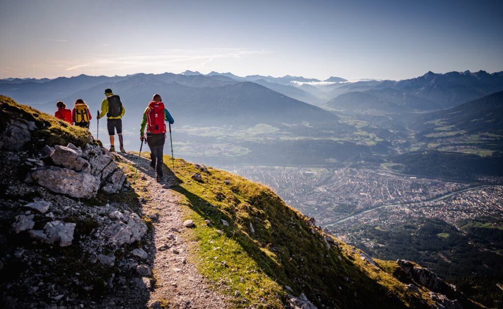 Vier Wanderer mit Rucksäcken gehen auf einem Bergpfad mit Blick auf eine Stadt und ein Tal, mit Bergketten in der Ferne unter einem klaren Himmel - eine inspirierende Szene für Lehrerfortbildung in der Natur.