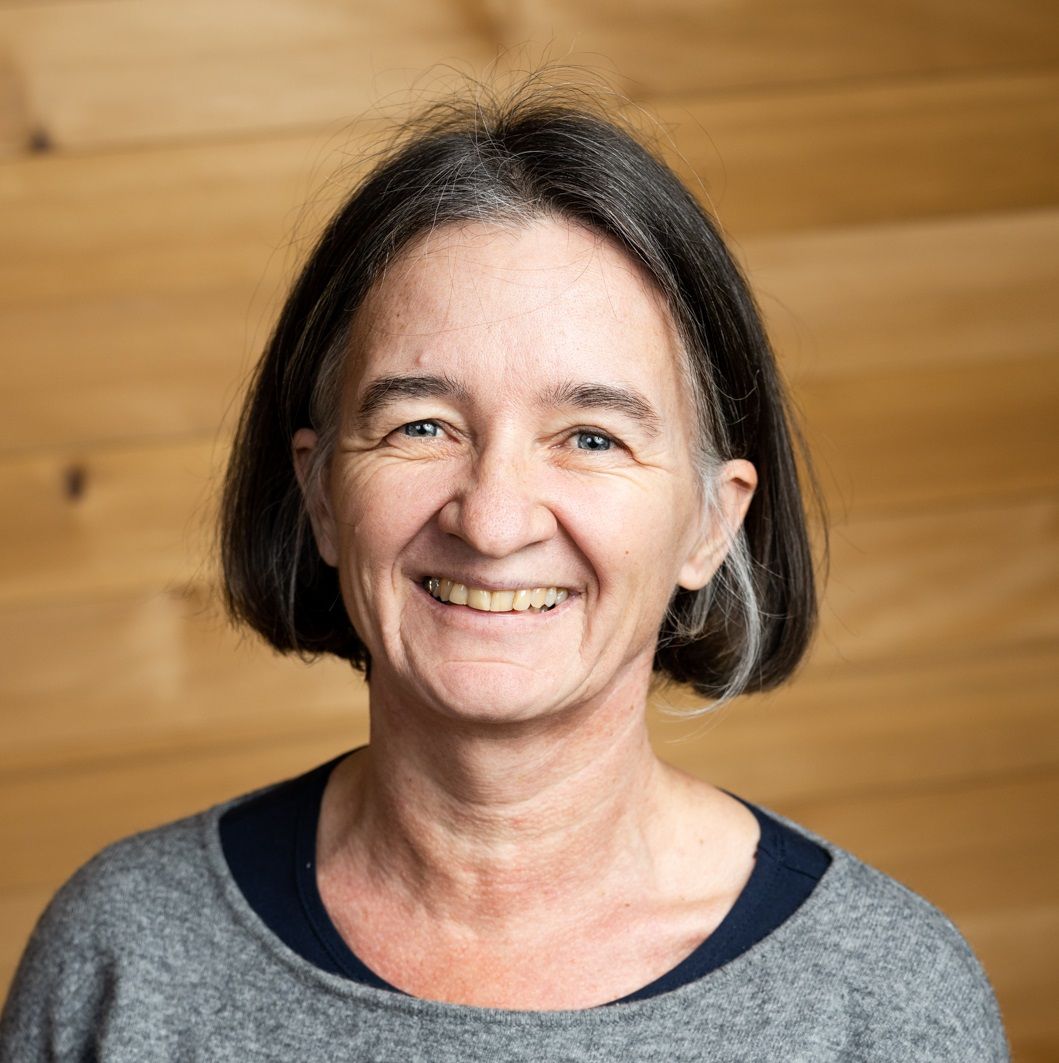 Valerie Braun, eine Frau mittleren Alters mit glattem, dunkelgrauem Haar und einem grauen Pullover, lächelt in die Kamera, während sie vor einer hellen Holzwand steht.