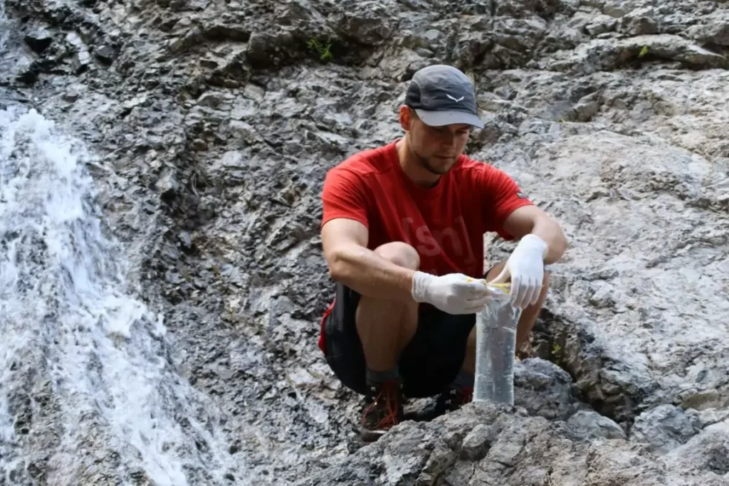 Eine Person mit Handschuhen und Mütze hockt auf einem Felsen in der Nähe eines Wasserfalls und sammelt im Rahmen der aktuellen Forschung eine Wasserprobe in einem durchsichtigen Plastikbeutel.