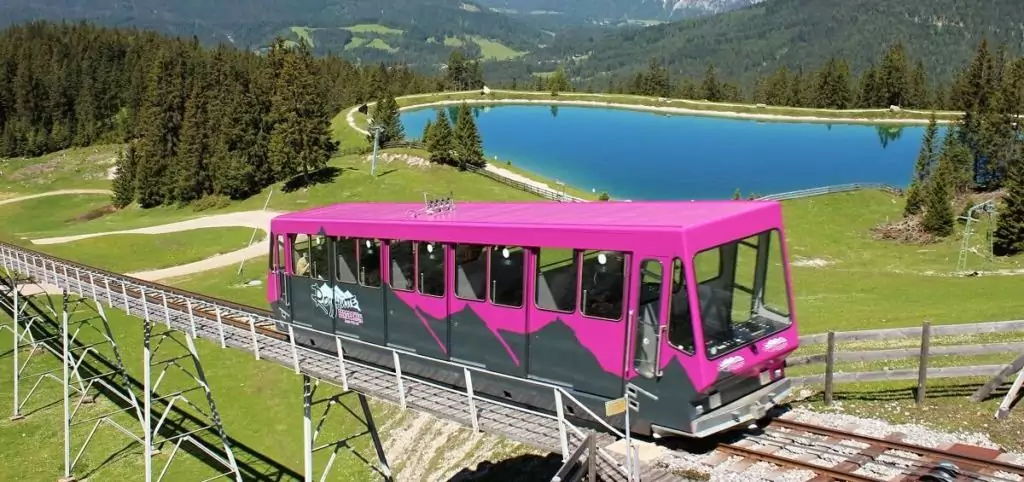 Ein rosafarbener Bergbahnwagen fährt einen Hang hinauf, umgeben von einem Waldgebiet, Gras und einem blauen Teich im Hintergrund.