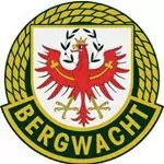 Kreisförmiges Emblem mit grüner und gelber Umrandung, das einen roten Adler mit Krone und Lorbeerkranz auf einem weißen Schild zeigt, der die stolze Partnerschaft der Organisation BERGWACHT" symbolisiert, die unten abgebildet ist.