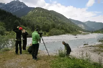 Vier Personen stehen an einem Fluss in einer bergigen Gegend; eine von ihnen hält mit einer Stativkamera Momente für die aktuelle Forschung fest, während eine andere in der Nähe des Wasserrandes steht. Im Hintergrund sind Bäume und Berggipfel zu sehen.