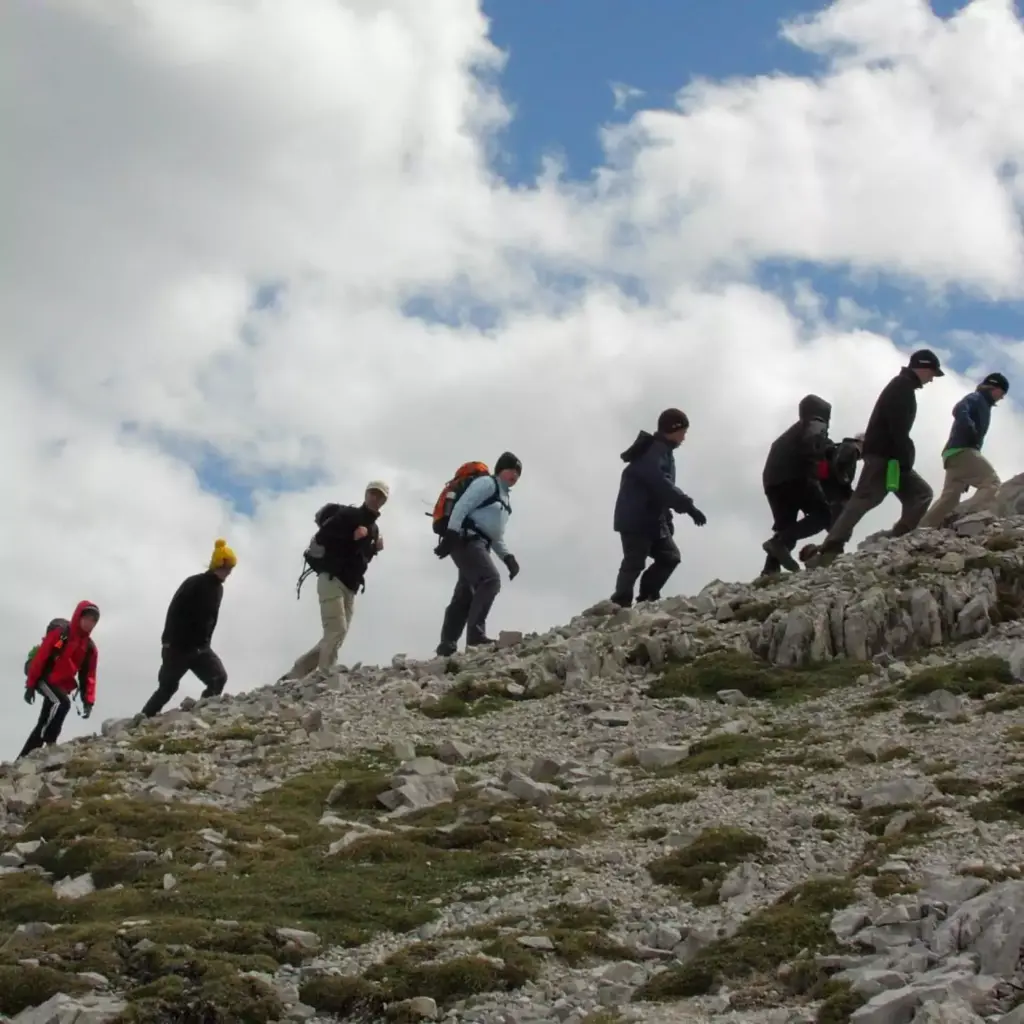 Eine Gruppe von Wanderern in Outdoor-Ausrüstung geht in einer Reihe auf einem felsigen, abschüssigen Bergpfad unter einem teilweise bewölkten Himmel während eines Expeditionsabenteuers im Karwendel.