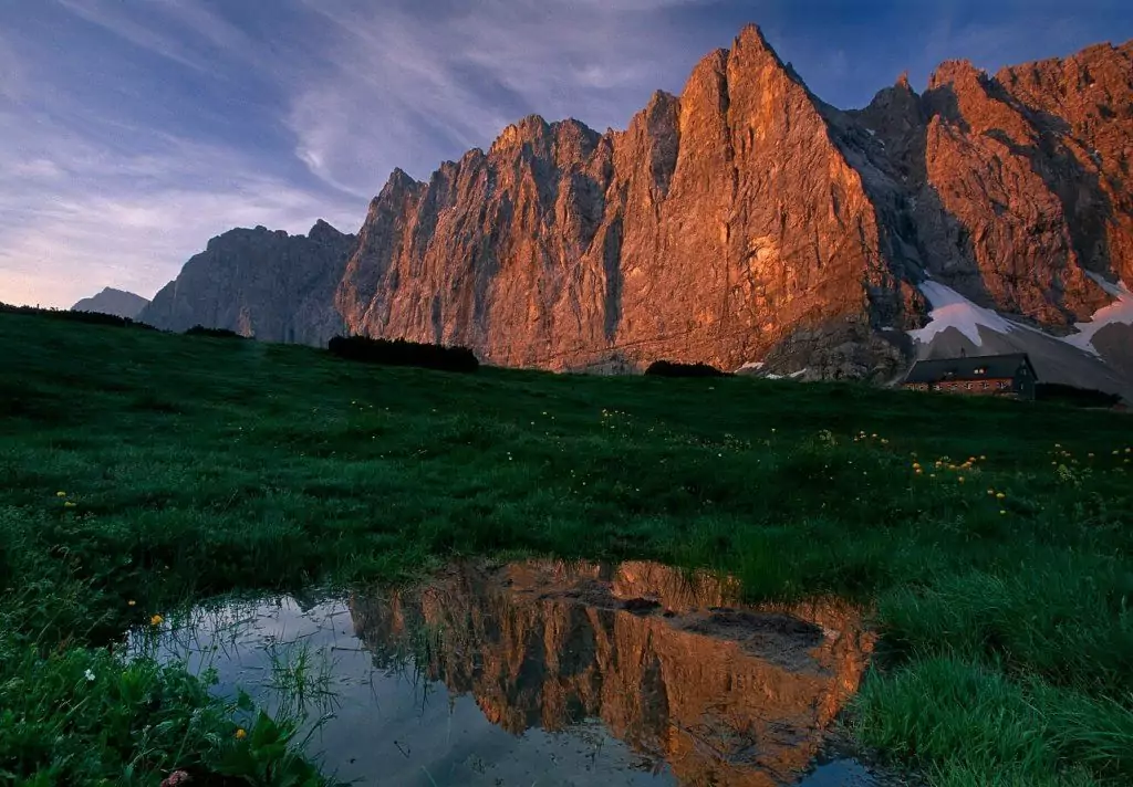 Felsige Bergklippen im Licht des Sonnenuntergangs mit grünem Gras im Vordergrund und einer kleinen Pfütze, in der sich der Berg spiegelt - eine inspirierende Szene, die an naturnahe Forschungsprojekte erinnert.