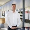Martin Friede, bekleidet mit einem weißen Hemd, steht drinnen an einem Tresen und lächelt in die Kamera. Im Hintergrund sind Regale und Dokumente zu sehen.