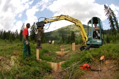 Eine Person bedient einen Bagger mit einem Bohrgerät, während eine andere in der Nähe in einem bewaldeten, bergigen Gebiet mit in den Boden eingelassenen Holzpfählen steht, die Natur- und Artenschutzprojekte unterstützen.