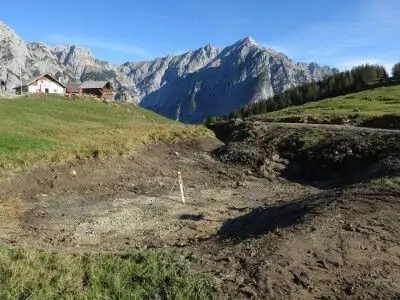 Ein grasbewachsener Hügel mit einem Fleck kahler Erde im Vordergrund, Häuser zur Linken und Berge im Hintergrund unter einem klaren blauen Himmel - eine ideale Kulisse für Natur- und Artenschutzprojekte.