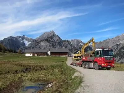 Ein roter Lastwagen transportiert einen gelben Bagger auf einer Landstraße mit Bergen, grasbewachsenen Feldern und landwirtschaftlichen Gebäuden im Hintergrund unter blauem Himmel - eine ideale Kulisse für Natur- und Artenschutzprojekte.