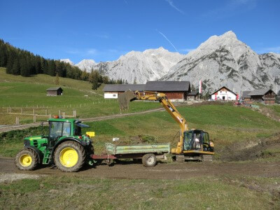Ein grüner Traktor mit Anhänger ist auf einem Feldweg geparkt, während ein gelber Bagger Material in den Anhänger lädt; Berge und landwirtschaftliche Gebäude im Hintergrund unterstreichen die Bedeutung von Natur- und Artenschutzprojekten in ländlichen Gebieten.