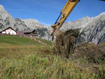 Ein Bagger hebt einen Erdbrocken auf einem grasbewachsenen Feld mit Bergen und Alpenhäusern im Hintergrund unter einem strahlend blauen Himmel aus und unterstützt damit Natur- und Artenschutzprojekte in dieser unberührten Landschaft.