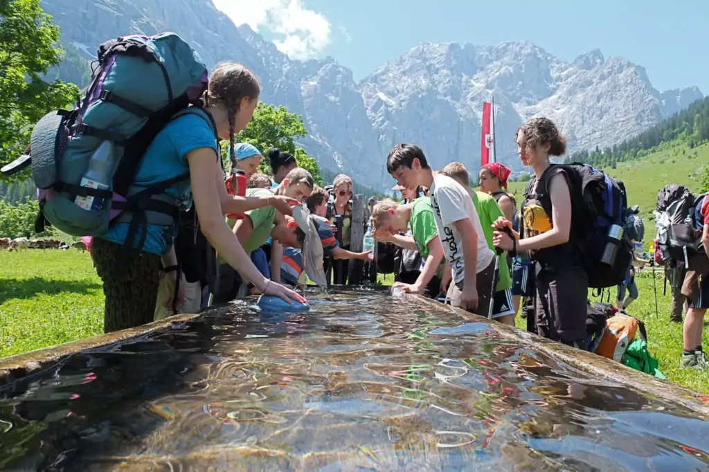 Eine Gruppe von Wanderern mit Rucksäcken, die an der Expedition Karwendel teilnimmt, versammelt sich um einen rechteckigen Wassertrog auf einer Wiese mit Bergen im Hintergrund.
