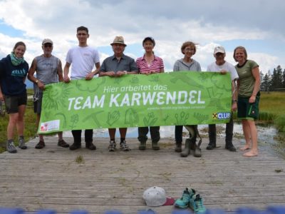 Neun Personen stehen im Freien auf einer hölzernen Plattform und halten ein grünes Transparent mit der Aufschrift "Hier arbeitet das Team Karwendel", das ihr Engagement für Natur- und Artenschutzprojekte symbolisiert. Auf dem Boden davor liegen Wanderschuhe und ein Hut.