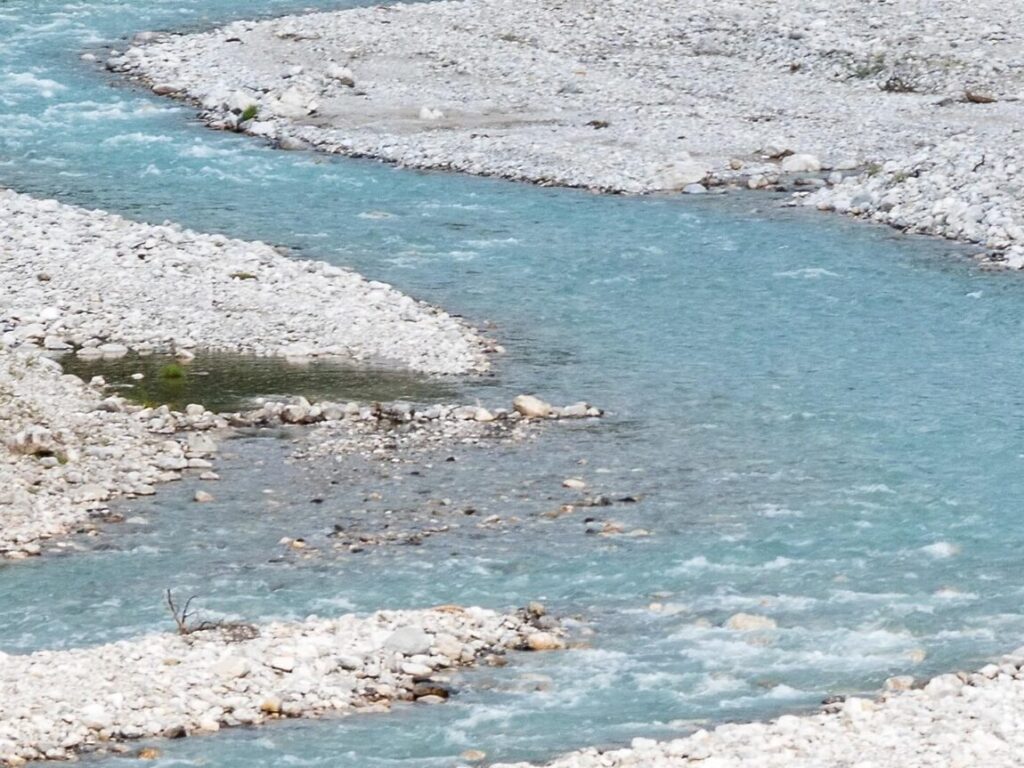 Ein flacher, gewundener Fluss mit klarem, blauem Wasser fließt über ein felsiges, kiesbedecktes Flussbett - eine ideale Kulisse für KlimaPädagogik-Aktivitäten, die die Bedeutung natürlicher Wasserwege hervorheben.