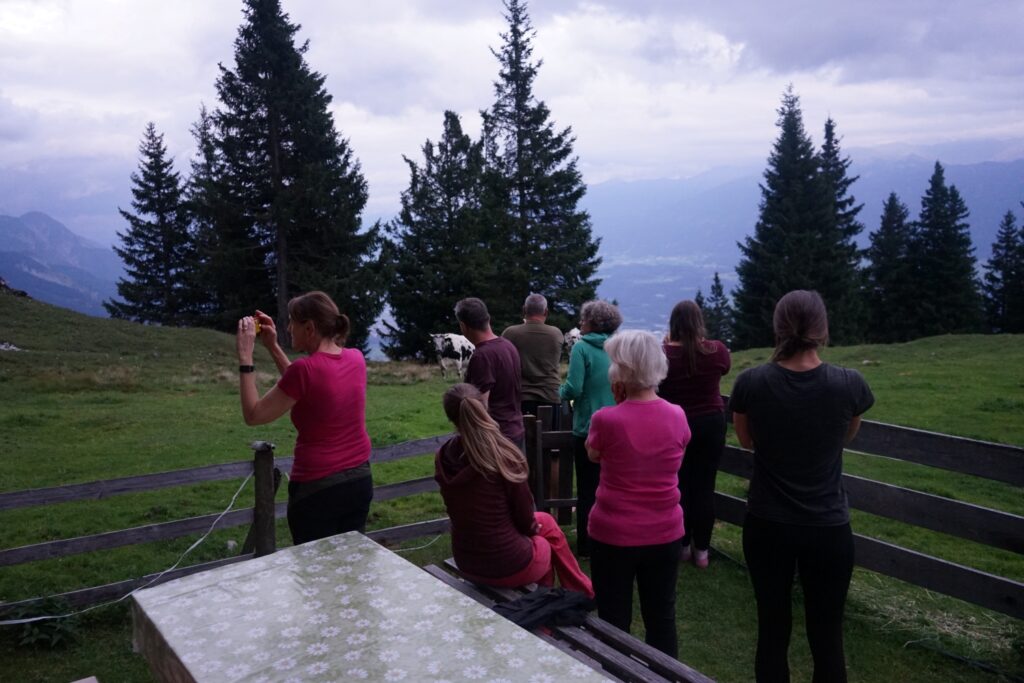 Eine Gruppe von Menschen steht und sitzt vor einer malerischen Bergkulisse in der Nähe von Zirbenbäumen auf der Achselkopfalm, mit einem Tisch im Vordergrund und bewölktem Himmel darüber.