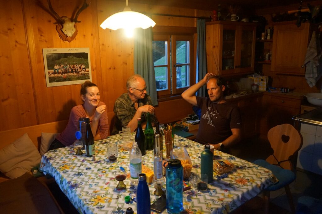 Drei Personen sitzen um einen Tisch mit Getränken und Snacks in einer gemütlichen, holzgetäfelten Küche auf der Achselkopfalm, beleuchtet von einer Hängelampe.