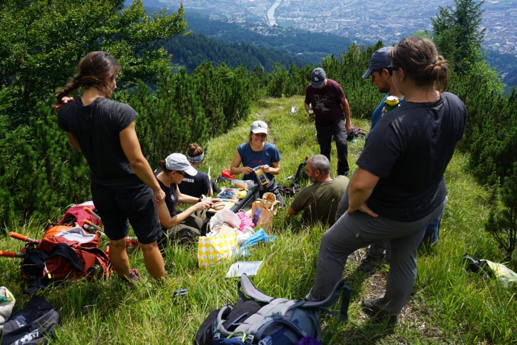 Eine Gruppe von Menschen sitzt und steht auf dem grasbewachsenen Hang der Achselkopfalm und macht ein Picknick mit Rucksäcken und Lebensmitteln, mit Blick auf die Stadt und den Fluss darunter.