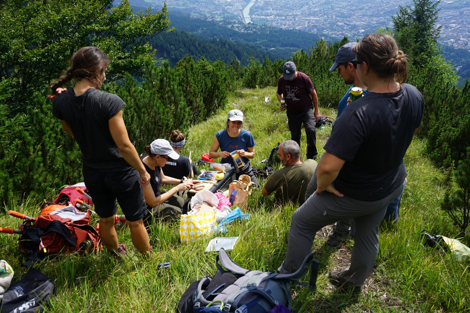 Eine Gruppe von Menschen sitzt und steht auf dem grasbewachsenen Hang der Achselkopfalm und macht ein Picknick mit Rucksäcken und Lebensmitteln, mit Blick auf die Stadt und den Fluss darunter.
