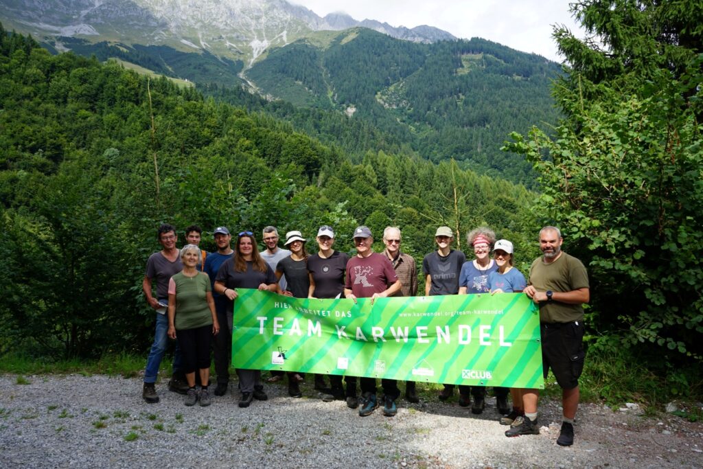 Eine Gruppe von Menschen steht im Freien vor einem bewaldeten Berg in der Nähe der Achselkopfalm und hält ein grünes Transparent mit der Aufschrift "Team Karwendel".