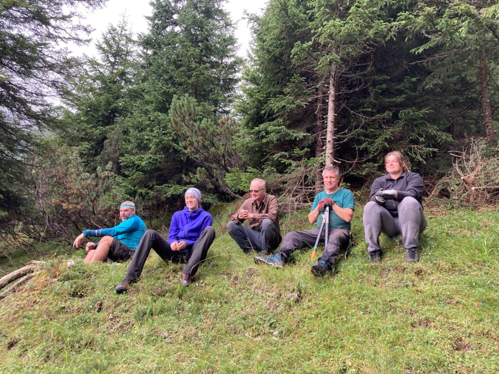 Fünf Personen sitzen auf einem grasbewachsenen Hang in der Nähe der Achselkopfalm vor Zirbenbäumen, in Outdoor-Kleidung, und scheinen bei einer Wanderung eine Pause zu machen.