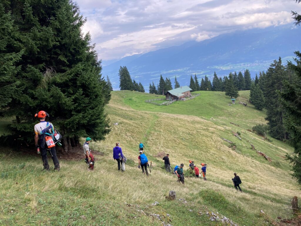 Eine Gruppe von Menschen in Outdoor-Ausrüstung wandert einen grasbewachsenen Hang hinunter zur Achselkopfalm, umgeben von Bäumen und Bergen unter einem bewölkten Himmel.