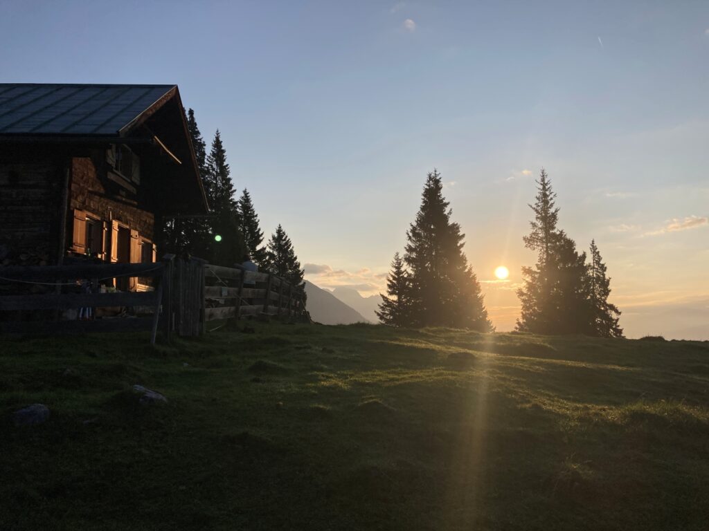 Eine hölzerne Hütte steht auf einem grasbewachsenen Hügel bei Sonnenuntergang. Das Sonnenlicht fällt durch hohe Kiefern in der Nähe der Achselkopfalm, im Hintergrund sind die majestätischen Berge zu sehen.