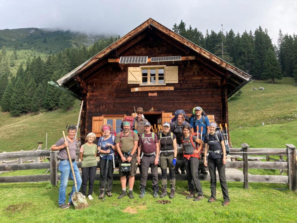 Eine Gruppe von Menschen mit Wanderausrüstung steht vor der Achselkopfalm, einer hölzernen Berghütte, die von Bäumen und grasbewachsenen Hügeln umgeben ist.