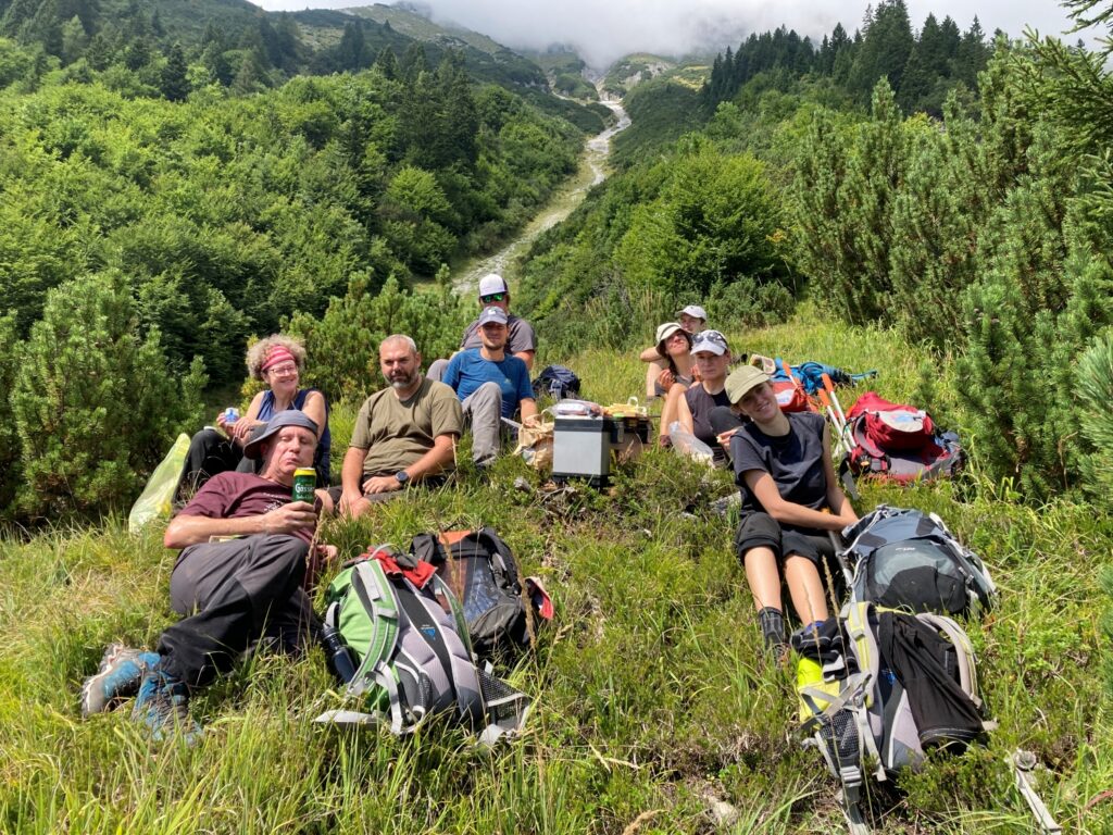 Eine Gruppe von Menschen ruht sich mit Rucksäcken und Outdoor-Ausrüstung auf einer grasbewachsenen Anhöhe bei der Achselkopfalm aus, umgeben von grünen Bäumen und Bergen im Hintergrund.