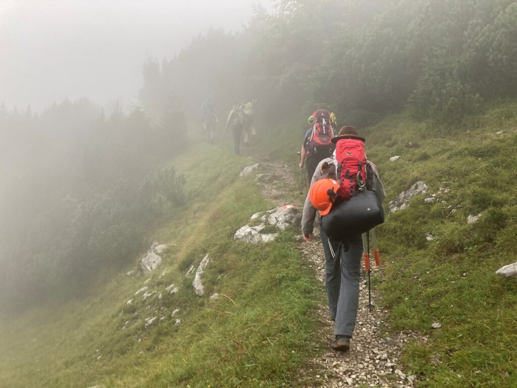 Eine Gruppe von Wanderern mit Rucksäcken geht im Gänsemarsch einen schmalen, felsigen Pfad durch nebliges, grasbewachsenes Gelände bei der Achselkopfalm hinauf.