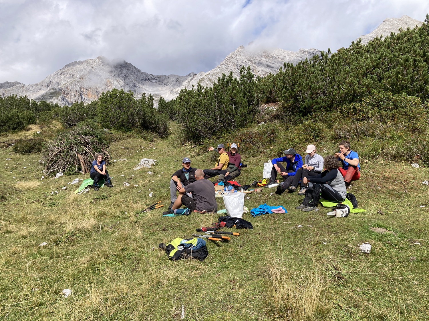 Eine Gruppe von Menschen sitzt im Gras auf einer Bergwiese bei der Almpflege Martinsbergalm und macht eine Pause, umgeben von Outdoor-Ausrüstung und Rucksäcken, mit Bergen und Sträuchern im Hintergrund.