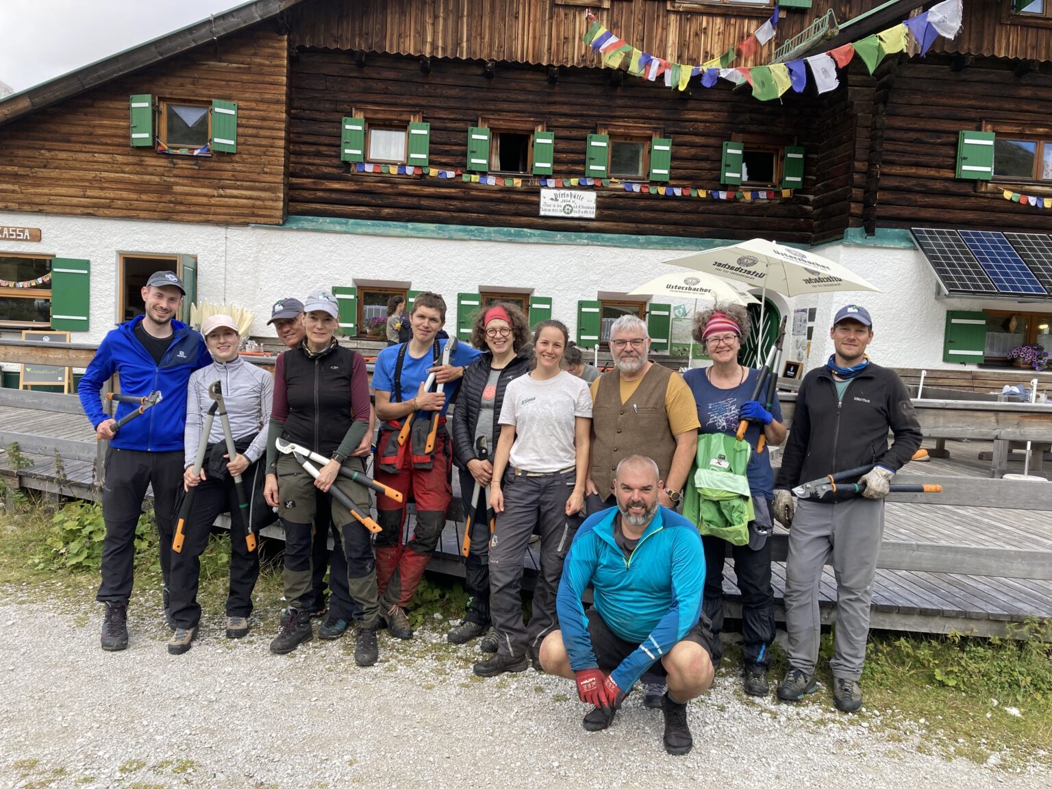 Eine Gruppe von elf Personen in Outdoor-Kleidung und -Ausrüstung posiert vor der Almpflege Martinsbergalm mit ihren grünen Fensterläden und bunten Gebetsfahnen.