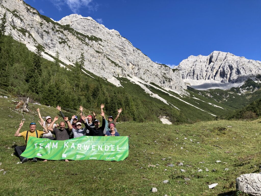 Eine Gruppe von Menschen hält im Freien ein grünes "Team Karwendel"-Banner hoch und reckt die Arme in die Höhe. Im Hintergrund sind die Berge und der strahlend blaue Himmel zu sehen, während einer Veranstaltung der Almpflege Martinsbergalm.