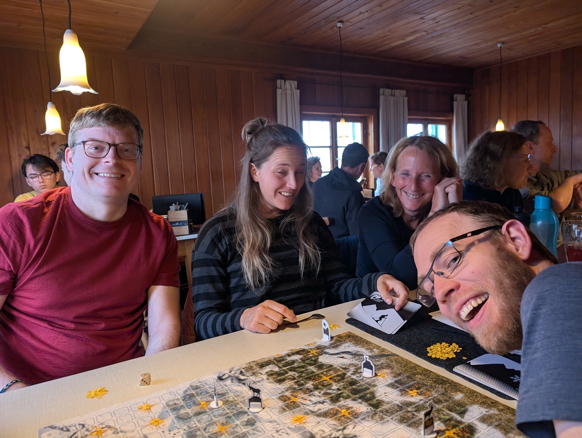 Vier Personen sitzen in einem holzgetäfelten Raum an einem Tisch und spielen ein Brettspiel. Sie lächeln und spielen mit Spielsteinen und Karten. Andere Personen sind im Hintergrund zu sehen.